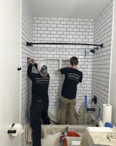 Two workers installing white subway tiles with black grout in a shower area, adding finishing touches.