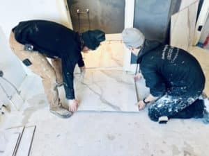 Two workers carefully placing a large marble-look tile on the floor during a bathroom installation.