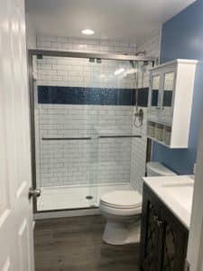 Bright bathroom with a white subway tile shower, accented by a bold blue tile stripe. Features sliding glass doors, wood-look flooring, and modern fixtures.